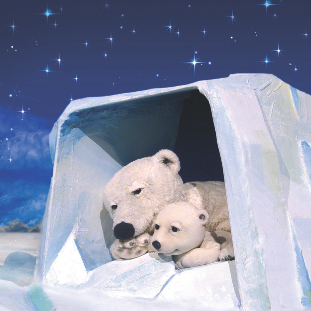 Zwei Eisbär-Puppen, ein großer und ein kleiner, liegen zusammen in einer gemütlichen Eishöhle unter einem sternklaren Nachthimmel – Szene aus dem Theaterstück ‚Der kleine Eisbär
