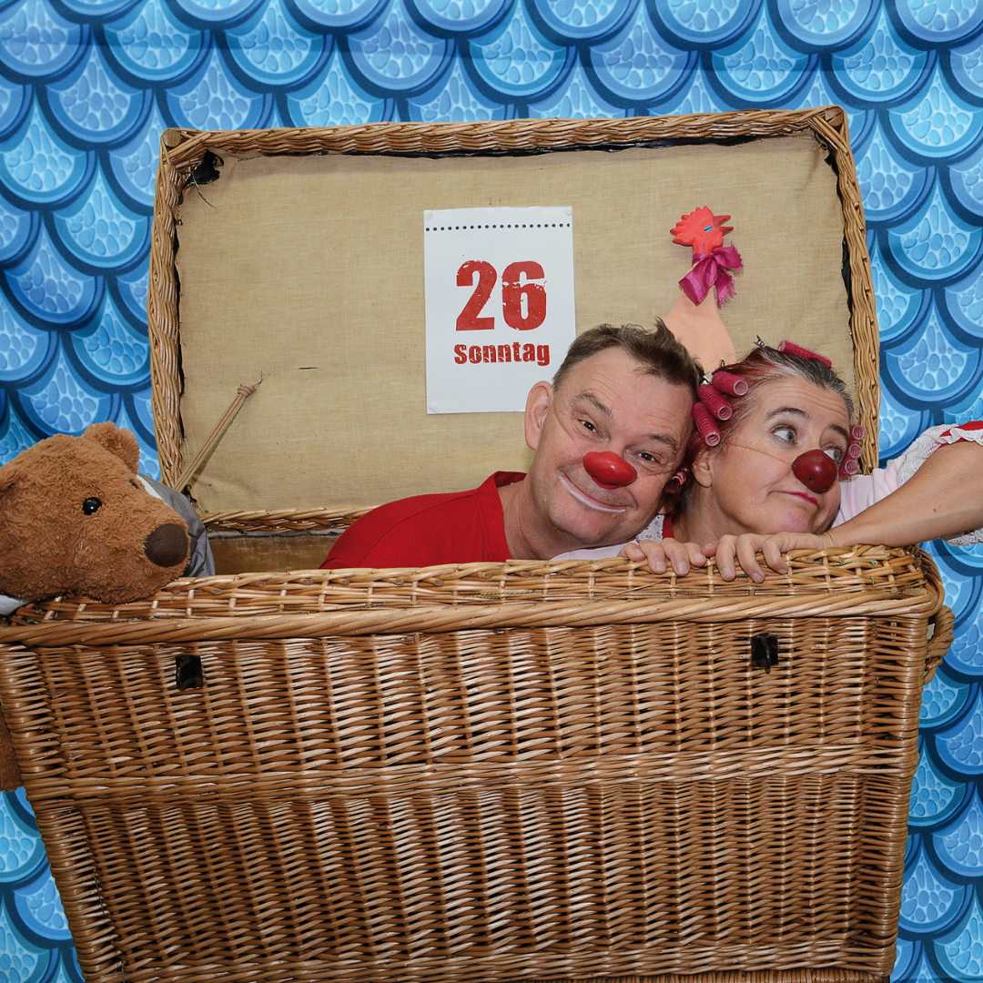 „Die Clowns Herbert und Mimi mit roten Nasen schauen fröhlich aus einer großen Wäschetruhe. Im Hintergrund ein blauer, schuppenartiger Vorhang und ein Kalenderblatt mit der Aufschrift ‚26 Sonntag‘. Neben ihnen ein Teddybär und ein Spielzeughuhn.