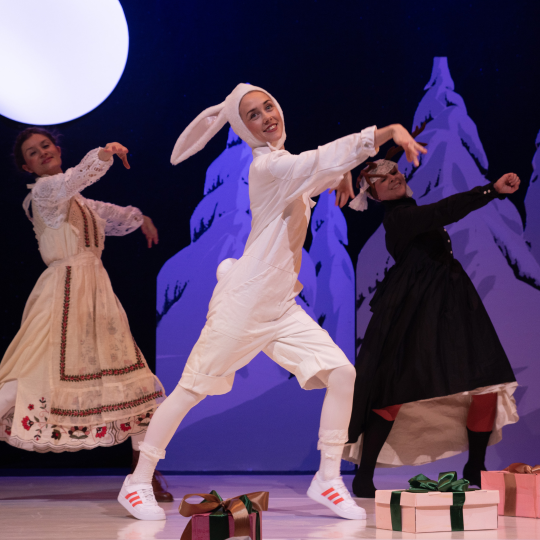 Drei Darstellerinnen tanzen auf der Bühne vor verschneiten Tannen, eine im weißen Hasenkostüm mit langen Ohren, daneben zwei weitere in folkloristischen Kleidern – Szene aus dem Theaterstück ‚Der Weihnachtshase‘.