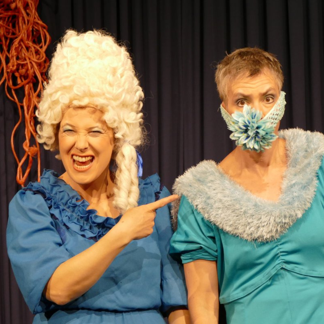 Zwei Schauspielerinnen in blauen Kostümen: eine mit weißer Lockenperücke lacht und zeigt auf die andere, die ernst blickt und eine große Blume als Mundmaske trägt. Szene aus dem Theaterstück ‚Knallwut‘.