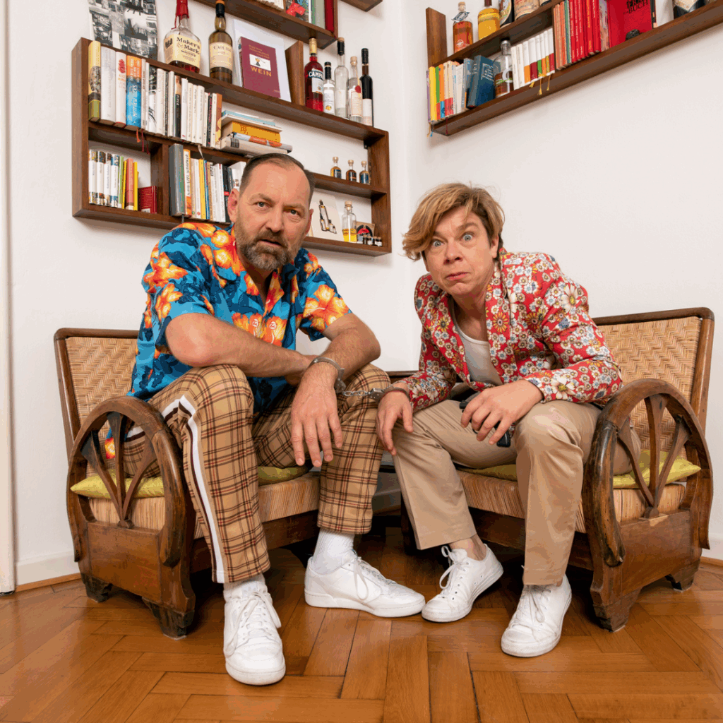 Birgit Linner und Roland Trescher vom Improtheater Isar 148 sitzen nebeneinander in bunten, gemusterten Outfits auf zwei Sesseln, blicken neugierig und konzentriert in die Kamera – eine humorvolle Szene, die ihre lebendige Improvisationskunst widerspiegelt.