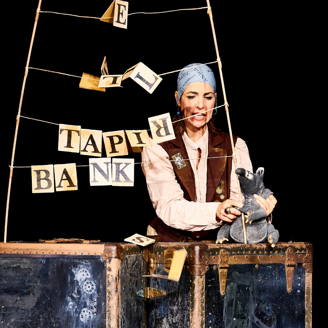 Szene aus dem Theaterstück ‚Pop-up Pirat‘: Eine Schauspielerin mit Kopftuch und Piratenweste steht hinter zwei alten Seemannskisten. In der Hand hält sie eine Tapir-Handpuppe, über ihr hängt eine schiefe Girlande mit den Buchstaben ‚TAPIR BANK‘.