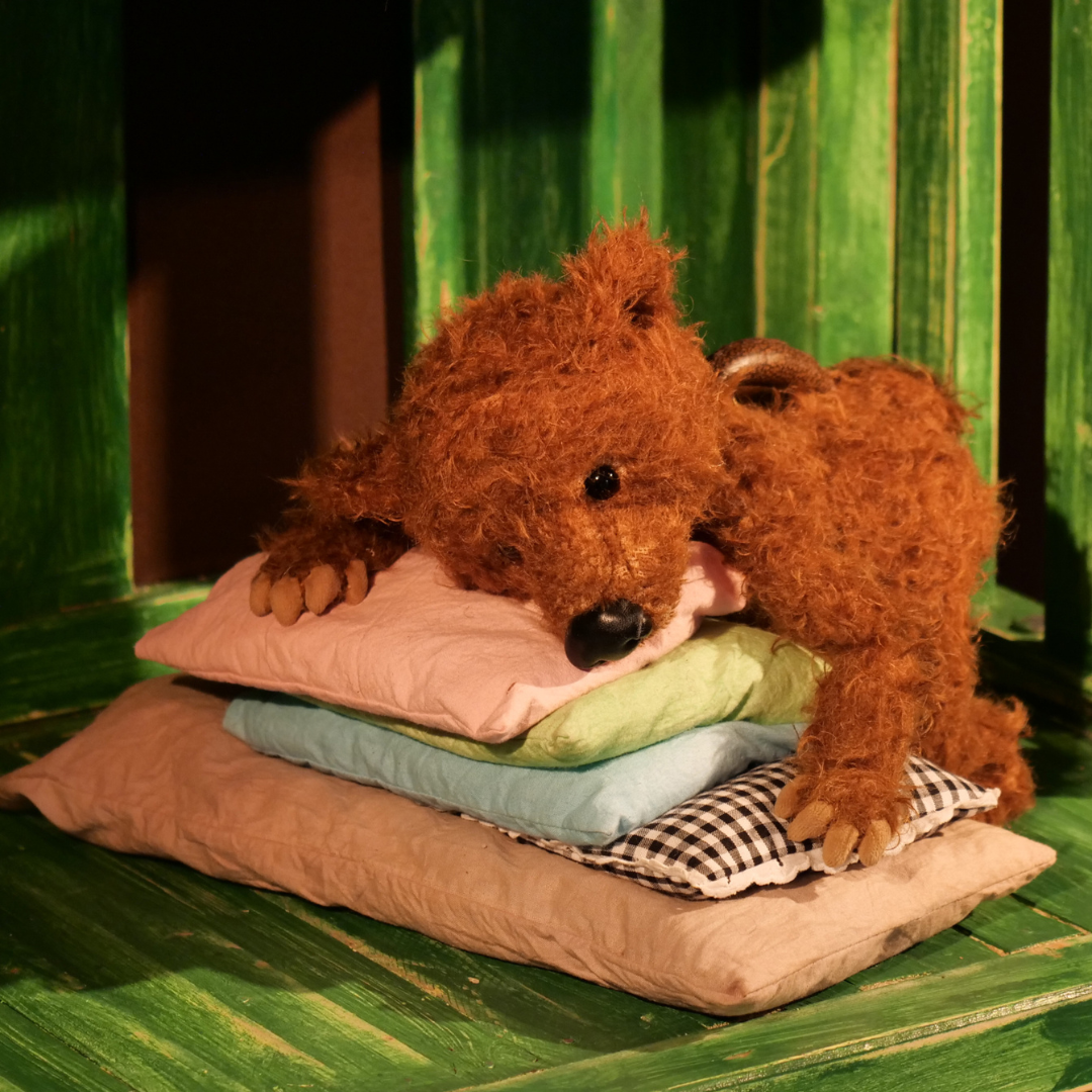Brauner Plüschbär liegt auf einem Stapel bunter Kissen in einer grünen Holzkulisse. Szene aus dem Kindertheaterstück ‚Bär hat es schwer‘.