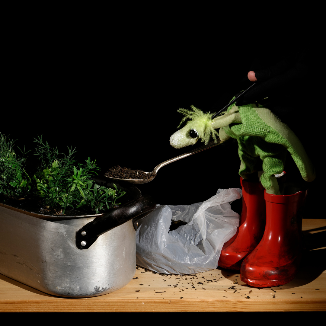 Grüne Handpuppe mit wuscheligem Kopf in roten Gummistiefeln hält einen Löffel mit Erde über einen Blumentopf mit Kräutern – Szene aus dem Figurentheaterstück ‚Als der kleine Koriander kam‘.