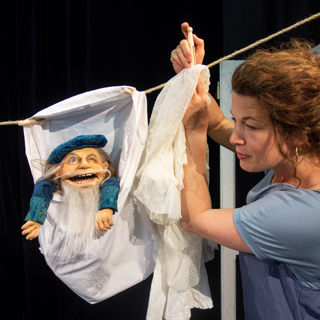 Eine Frau mit roten Haaren hängt eine weiße Stoffpuppe – ein Zwerg mit langem weißen Bart und blauer Mütze – an eine Wäscheleine. Szene aus dem Theaterstück ‚Neeweißnicht und Rosenrot‘.