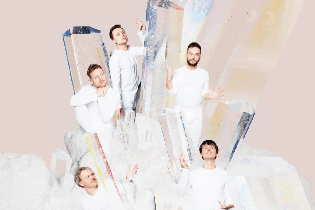 A-cappella-Gruppe Gesangskapelle Hermann posiert in weißen Outfits zwischen großen, irisierenden Kristallen vor einem pastellrosa Hintergrund.