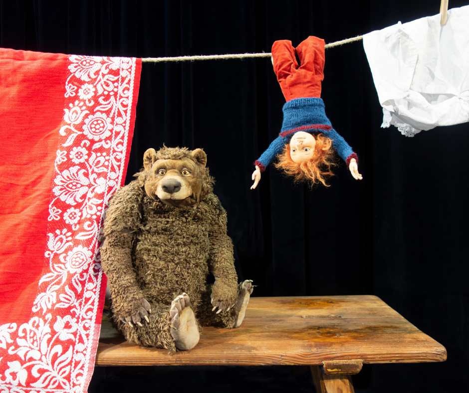 Bühnenfoto aus dem Kindertheaterstück „Neeweißnicht und Rosenrot“: Ein großer, freundlich wirkender Bärenfigur sitzt auf einem Tisch. Daneben hängt kopfüber eine Kinderpuppe mit roten Haaren an einer Wäscheleine, zwischen aufgehängten Tüchern.