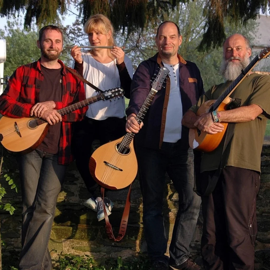 Vier Musiker der Band HüSCH! stehen im Freien vor Bäumen und einer Steinmauer. Drei von ihnen halten Saiteninstrumente (Mandoline und Gitarren) in den Händen, eine Musikerin steht in der Mitte. Die Gruppe blickt ruhig in die Kamera, die Atmosphäre wirkt natürlich und bodenständig.