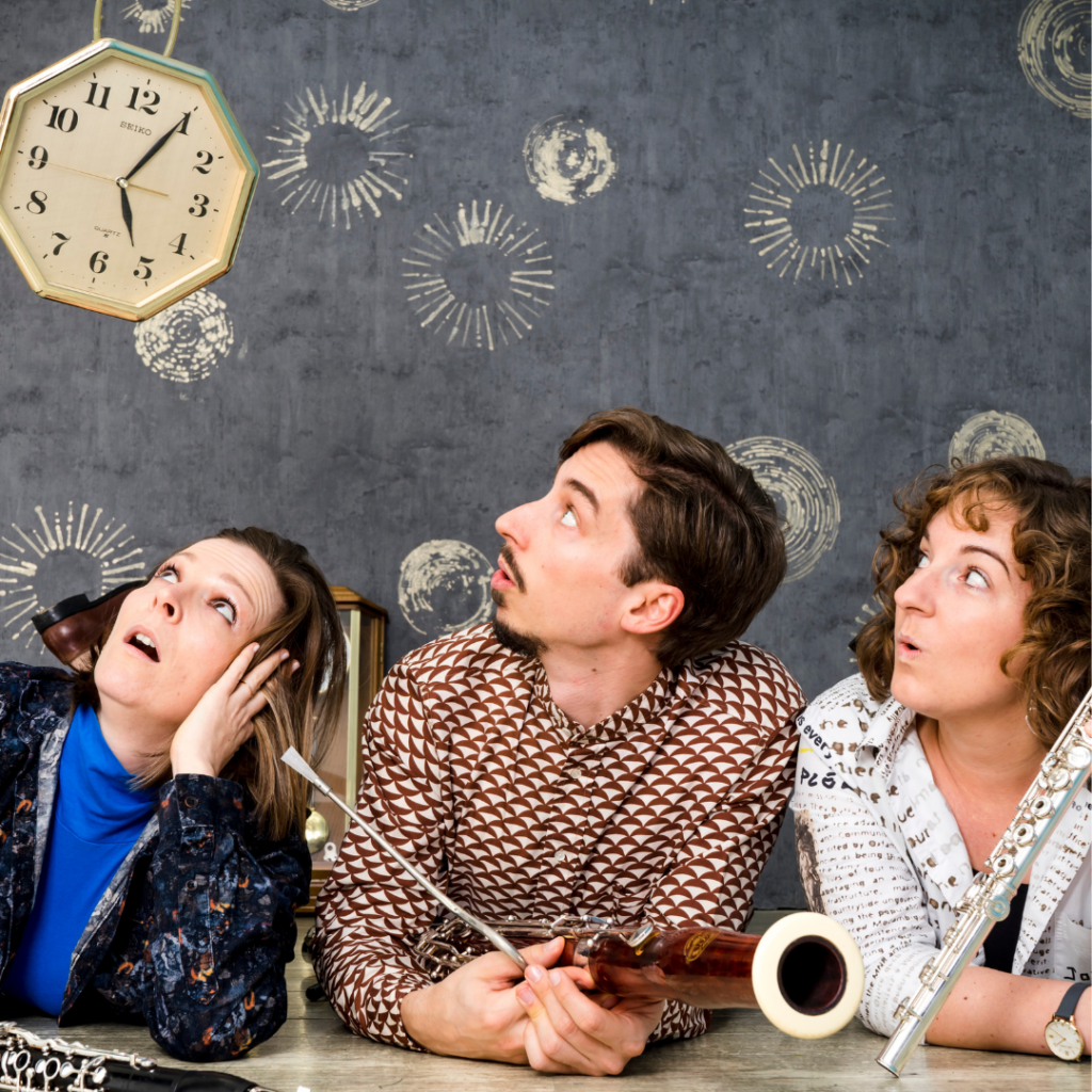 Drei Mitglieder des Ensembles Blasbalg sitzen mit Flöte, Klarinette und Fagott an einem Tisch und blicken überrascht nach oben zu einer Wanduhr. Im Hintergrund ist eine dunkelgraue Wand mit grafischen Kreis- und Zeitmotiven zu sehen.