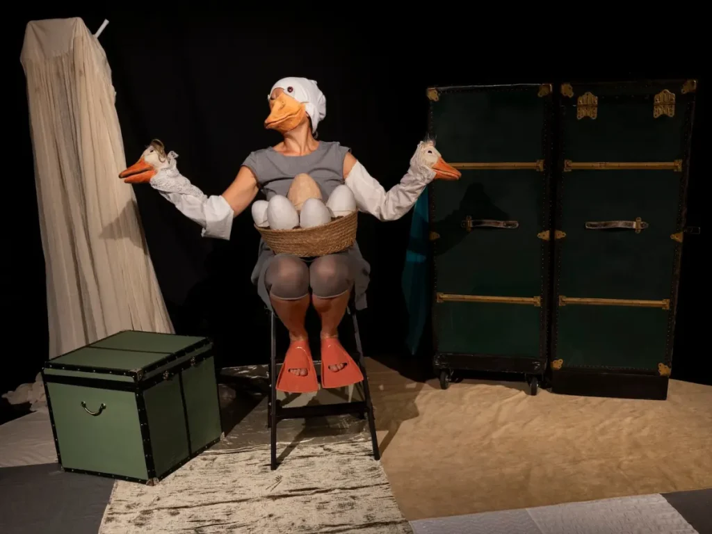 Szene aus dem Kindertheaterstück „Schwanen“: Eine Darstellerin mit Entenkopf und Handpuppen sitzt mit einem Korb voller Eier auf der Bühne, umgeben von großen grünen Koffern.