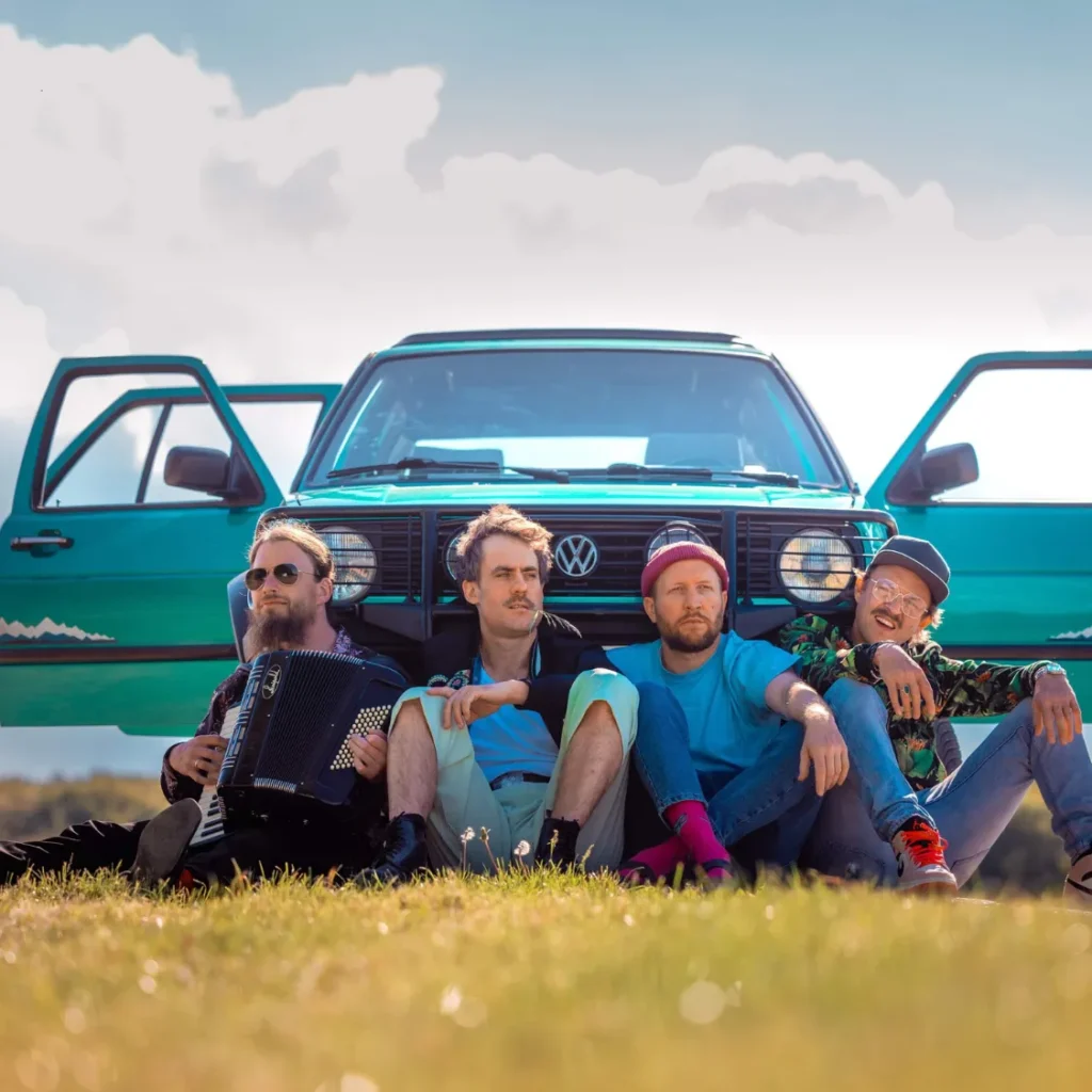 Vier Musiker von Gankino Circus sitzen mit einem Akkordeon auf einer Wiese vor einem türkisfarbenen VW-Bus mit geöffneten Türen.